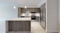 Located at 4200 NW 107 Ave in Doral, FL, this well-appointed single-floor unit offers a thoughtfully designed layout with modern finishes throughout. The kitchen is a standout feature, showcasing wood-tone cabinetry, stainless steel appliances including a gas range, built-in microwave, and French door refrigerator, all complemented by a geometric hexagonal tile backsplash and crisp white quartz countertops. A large island with pendant lighting above provides additional prep and dining space, while track lighting illuminates the full length of the kitchen. Light wood-style flooring flows seamlessly throughout the unit, creating a bright and cohesive aesthetic. The living and bedroom areas are generously proportioned with neutral-toned walls that maximize the sense of openness. Bedrooms feature natural light from windows with views to the outside. The bathroom is finished with classic white subway tile in a spacious walk-in shower enclosed by a frameless glass door, paired with recessed ceiling lighting. A dedicated laundry area houses a stacked Whirlpool washer and dryer unit for added convenience. Clean lines, neutral tones, and contemporary fixtures define the overall ambiance of this modern Doral residence.