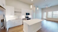 This 1,070-square-foot single-story residence offers 3 bedrooms, 2 bathrooms, and 1 closet, thoughtfully laid out for both comfort and functionality. The open-concept kitchen and living area serves as the heart of the home, showcasing soaring ceilings, pendant lighting, and a large central island with crisp white quartz countertops. White shaker-style cabinetry extends to the ceiling, complemented by stainless steel appliances including a French door refrigerator, electric range, microwave, and dishwasher. Open shelving and a tile backsplash add refined detail. Light hardwood flooring runs consistently throughout the entire unit, lending warmth and cohesion. The living area features tall ceilings, a ceiling fan, and a glass door that opens to a balcony with exterior views, allowing ample natural light. Each of the three bedrooms is finished with the same hardwood flooring, neutral walls, and window blinds, with ceiling fans in select rooms. The primary bedroom measures 170 square feet and connects to a dedicated closet and a private bathroom corridor. Both bathrooms are finished in white with clean vanities, white cabinetry, and wood-look flooring. The overall aesthetic is modern, clean, and move-in ready.
