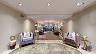 This sophisticated single-floor space presents a masterfully designed lobby and common area that exudes classical elegance at every turn. The grand entrance hallway sets the tone immediately, featuring an ornate medallion-patterned carpet, symmetrical tufted velvet seating in muted purple, gilded wall sconces, and large framed mirrors that amplify the sense of depth and grandeur. A chandelier overhead adds a refined focal point as the corridor leads deeper into the space.

Beyond the entry corridor, a generously proportioned lounge area opens up, anchored by a striking grand piano and ringed by plush velvet sofas and armchairs arranged around glass and gold coffee tables. Wainscoting and decorative panel molding line the walls, while patterned carpeting in soft sage green adds warmth underfoot. A structural column is seamlessly integrated into the layout, and wall-mounted sconces provide a soft, ambient glow.

The main entrance foyer features an intricate inlaid tile floor, travertine-flanked staircase risers adorned with floral arrangements, and glass entry doors that invite natural light deep into the space. A separate, well-lit office area provides a functional counterpoint, equipped with a classic wooden desk, mailboxes, a corkboard, and sheer-draped windows that fill the room with diffused natural light. Together, these spaces form a cohesive, upscale environment of timeless character.