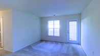 This single-floor residence offers a clean, straightforward layout designed for comfortable everyday living. The main living area is a standout feature, characterized by soft carpet flooring and neutral-toned walls that create a calm and versatile backdrop suitable for a variety of interior styles. A large multi-pane window serves as the focal point of the room, flooding the space with generous natural light and contributing to an open, airy atmosphere. Adjacent to the window, a glass-panel exterior door with slatted blinds provides both additional light and convenient access to the outdoors. The walls are smooth and well-maintained, complemented by clean white baseboards that frame the carpeted floor. The ceiling is finished with a subtle texture and includes a central ventilation vent, reflecting a practical and functional design approach throughout. The overall ambiance of the space is bright, neutral, and move-in ready, making it an adaptable environment well-suited for a range of furnishing and decorating preferences. The single-level layout ensures ease of navigation throughout, and the abundance of natural light remains a defining characteristic of the home's interior character.