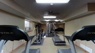 This fitness center is a dedicated, single-floor exercise facility designed to support a comprehensive workout routine. The room is laid out in a long, open floor plan that comfortably accommodates multiple pieces of equipment without feeling cramped. Cardio machines, including treadmills and an elliptical trainer, are arranged along the central and peripheral areas of the room, providing ample space for users to move freely between stations. A multi-station strength training machine anchors the middle of the space, offering a range of resistance-based exercises. The flooring consists of a combination of carpet and protective rubber mats beneath the equipment, ensuring both comfort and safety. Overhead, a ceiling fan provides air circulation, complementing the fluorescent lighting that keeps the space well-lit throughout. A skylight panel further enhances the brightness of the room. Natural light filters in through windows positioned along one wall, adding to the open and energizing atmosphere. The neutral, warm-toned walls and low-profile ceiling contribute to a functional and focused environment well-suited for fitness activities. The overall ambiance is clean, practical, and purposefully arranged to serve users looking for a well-rounded exercise experience.