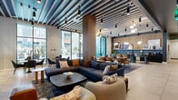 Located at 39176 Fremont Blvd in Fremont, California, this thoughtfully designed single-floor amenity space offers a sophisticated and functional environment for gathering, working, and relaxing. The layout flows seamlessly across its generously sized footprint, encompassing a living room and dining area complemented by several distinct zones.

The entrance corridor features vertical wood slat partitions and large-format porcelain tile flooring, setting a contemporary tone. The living area is anchored by a plush sectional sofa and accent chairs arranged around a wall-mounted flat-screen display, with a striking vertical slat divider adding architectural depth and visual interest.

The dining and lounge zone is highlighted by floor-to-ceiling glass walls and sliding doors that flood the space with natural light and offer direct views of an outdoor courtyard and pool area. A boldly designed ceiling with alternating wood and painted beams adds dramatic character overhead, while pendant globe lights and track lighting provide layered illumination.

A built-in coffee and refreshment bar features rich wood cabinetry, deep blue tile backsplash, and a long counter with bar seating — ideal for casual meetings or morning coffee. A dedicated conference room with a wood table, executive chairs, and a wall-mounted display provides a private setting for focused work or small meetings. The overall ambiance is modern, warm, and inviting throughout.