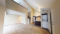 This two-story space makes an immediate impression with its dramatic vaulted ceilings that soar overhead in the main living area, creating a sense of openness and volume rarely found in comparable layouts. The living area is finished with wall-to-wall neutral carpeting, contributing to a warm and comfortable atmosphere, while light-toned walls reflect ambient light throughout the space.

A staircase leads to the upper level, which overlooks the main living area through an open loft-style railing, reinforcing the vertical dimension of the space. The kitchen is positioned adjacent to the living area in an open-concept arrangement, featuring dark espresso cabinetry with under-cabinet lighting, granite-style countertops, a stainless steel refrigerator, and a pendant light fixture above the counter. Track lighting provides additional illumination across the kitchen workspace.

Tile flooring anchors the kitchen and transition zones, offering a durable contrast to the carpeted living space. A dedicated laundry area with a stacked washer and dryer unit is conveniently located just off the kitchen, adding practical functionality. An additional room, accessible through a doorway near the living area, provides flexible usage potential.