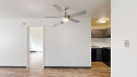 This single-floor unit offers a practical and well-laid-out living space finished with warm hardwood-style flooring that runs throughout the entirety of the home, creating a cohesive and inviting atmosphere.

The main living area is open and bright, centered beneath a brushed nickel ceiling fan with an integrated light fixture. The space flows naturally into the kitchen, which is set apart by a partial dividing wall. The kitchen features dark espresso-toned cabinetry, black appliances including a refrigerator and electric range, and a sink positioned along the counter. Recessed and flush-mount lighting keeps the kitchen well illuminated.

A dedicated bedroom provides a comfortable retreat, featuring a large window fitted with vertical blinds that allow ample natural light to filter in. A baseboard heater and a wall-mounted air conditioning unit ensure year-round climate control. The room offers generous wall space, providing flexibility for furniture arrangement.

The bathroom includes a wide vanity with a full-width mirror above a clean white countertop and drop-in sink with chrome fixtures.

Throughout the unit, crisp white walls with dark baseboards provide a clean, neutral backdrop. A hallway connects the main areas, offering additional storage access. The overall layout is efficient, functional, and move-in ready.