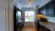 This single-story space showcases a well-appointed galley-style kitchen designed for efficient use of space. The kitchen features dark espresso cabinetry lining both sides of the corridor layout, creating a striking contrast against the light-colored walls and warm hardwood-style flooring. Stainless steel appliances, including a full-size refrigerator and a gas range with overhead ventilation hood, provide a sleek, modern aesthetic. The granite-style countertops complement the dark cabinetry, adding a touch of elegance to the functional layout. A centrally positioned window at the end of the kitchen draws in natural light, brightening the space and offering a pleasant outdoor view of greenery. Track lighting mounted on the ceiling provides additional illumination, ensuring the kitchen is well-lit throughout the day and evening. The neutral white walls keep the space feeling open despite the linear layout. The hardwood-style flooring runs throughout, adding warmth and continuity to the overall design. The kitchen's layout maximizes storage and workspace, making it both practical and visually appealing.