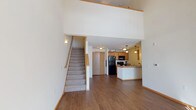 This two-story residence is defined by its striking vaulted ceilings in the main living area, creating an expansive, airy atmosphere that immediately draws the eye upward. The main level features warm hardwood-style flooring that runs throughout the open-concept living and dining areas, lending a clean and cohesive look to the space. A carpeted staircase with wood railings connects the two floors seamlessly.

The kitchen is well-appointed with warm honey-toned cabinetry extending to the ceiling for ample storage, a peninsula countertop ideal for casual dining or meal prep, and a full suite of black appliances including a refrigerator, range, and microwave. Pendant lighting above the peninsula adds a functional yet stylish touch.

The upper level offers a versatile open loft space overlooking the main floor below, carpeted for comfort and outfitted with a ceiling fan. Additional rooms on this level include a carpeted bedroom with a ceiling fan and a window providing natural light, as well as two bathrooms — one featuring a double-sink vanity and a glass-enclosed walk-in shower, and another with a tub and shower combination. A dedicated laundry area with a full-size washer and dryer, plus overhead shelving, adds everyday convenience to the home.
