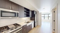 Located at 514 SE Belmont St. in Portland's vibrant SE neighborhood, this modern single-floor unit offers a thoughtfully designed layout comprising a living area, kitchen, and bathroom. The interior features light-toned hardwood flooring throughout, crisp white walls, and clean-lined architecture that creates a bright, open ambiance. The galley-style kitchen is a standout feature, equipped with full stainless steel appliances including a gas range, built-in microwave, dishwasher, and refrigerator. Dark espresso-toned cabinetry with sleek hardware contrasts elegantly against light quartz countertops and a classic subway tile backsplash. Open shelving adds both functionality and visual interest to the kitchen design. The living area is bathed in natural light from a large, black-framed picture window that frames views of the Portland skyline and surrounding greenery, lending an airy, elevated feel to the space. In-unit washer and dryer hookups provide added convenience. The bathroom features a full tub-shower combination with clean white tile surround. The overall design aesthetic is contemporary and minimalist, making excellent use of every square foot in this efficiently planned urban residence.