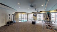 This fitness center in Houston, TX offers a thoughtfully designed, single-floor layout that accommodates a wide range of workout activities. The space is divided into distinct functional zones, beginning with a dedicated free weights area equipped with a full rack of dumbbells, medicine balls, barbells, resistance bands, and a cable machine — all reflected by floor-to-ceiling mirrors that enhance the sense of depth and support proper form during training.

The open exercise floor is generously proportioned, providing ample room for stretching, mat work, and functional training. A plyo box and yoga mat are positioned centrally, while a wall-mounted flat-screen television keeps users entertained. The space is finished with patterned carpet flooring and features crown molding with a tray ceiling detail, lending a polished aesthetic.

Large windows and glass doors line the perimeter, flooding the interior with natural light and offering direct views of the outdoor pool area below. A curved iron railing borders the pool-facing side, adding an architectural accent. Multiple ceiling fans and recessed lighting ensure consistent comfort and visibility throughout. The overall ambiance is bright, open, and well-appointed for a comprehensive fitness experience.