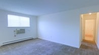 This single-floor space presents a clean, minimalist interior defined by bright white walls and neutral-toned wall-to-wall carpeting that lends a quiet, understated character to the room. The open layout offers a sense of spaciousness, with smooth white ceilings and simple, unadorned walls that provide a blank canvas adaptable to a variety of uses and furnishing styles.

Natural light enters through a window fitted with horizontal blinds, allowing for adjustable privacy and light control. Positioned beneath the window is a window-mounted air conditioning unit, ensuring cooling comfort during warmer months. Along the base of the wall, a baseboard heating unit provides an efficient and consistent heat source throughout cooler seasons, together forming a practical year-round climate control system.

The room's flooring consists of wall-to-wall carpeting in a muted, neutral tone, contributing to a soft and comfortable underfoot experience. A single electrical outlet is visible along one wall, reflecting a straightforward and functional design. Overall, the space maintains a simple, utilitarian aesthetic with clean lines, offering a versatile and move-in-ready environment.