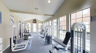 This fitness center offers a comprehensive workout environment designed to support a wide range of exercise routines. The space is defined by its striking vaulted ceilings, which lend an open, airy feel and contribute to a motivating atmosphere. A ceiling fan is mounted at the peak, ensuring comfortable airflow throughout the room. Large, multi-pane windows line the walls on multiple sides, flooding the interior with natural light and providing pleasant views of the lush greenery and brick exterior surroundings outside. The floor is finished with durable, cushioned fitness flooring, providing a safe and supportive surface for workouts. The room is outfitted with an array of strength training machines, including cable and weight stack systems, adjustable benches, and resistance equipment, as well as cardiovascular machines to round out the available options. A wall-mounted television is positioned at the far end of the room, visible from most workout stations. Double glass-paned doors at the rear offer both accessibility and additional light. Recessed lighting supplements the natural illumination, ensuring the space remains well-lit at all hours. Overall, the fitness center combines functionality with a bright, welcoming design that encourages regular use.