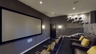 This dedicated screening room is designed to replicate the ambiance of a high-end private cinema. The room is arranged with two tiered rows of dark leather reclining chairs, each accented with gold velvet throw pillows, ensuring comfortable sightlines to the large projection screen mounted on the front wall. The screen is framed in black, commanding attention against the charcoal-toned walls. Recessed ceiling lighting and sleek wall sconces provide ambient illumination, while a ceiling-mounted projector delivers the visual presentation.

The rear of the room is elevated and features a stone-clad half-wall with a built-in counter, ideal for refreshments or entertaining. Chrome-railed stairs connect the two seating levels. Decorative spherical metallic wall sculptures and framed artwork add artistic character to the space. Floor-to-ceiling gold drapery lines one wall, adding warmth and acoustic value while lending a theatrical elegance. Deep blue patterned carpet covers the floors throughout, completing the sophisticated, cinema-inspired aesthetic. The overall design balances functionality with refined, upscale décor.