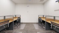 Located at 46120 Thomas Rd in Chilliwack, BC, this efficient office workspace offers a clean, modern environment designed to support focused, professional work. The single-room layout is thoughtfully organized with multiple individual workstations arranged along the perimeter and interior of the space. Each station features a warm-toned natural wood desk surface paired with sleek grey partition panels that provide a degree of privacy between users. Ergonomic mesh-back office chairs with chrome frames accompany each desk, contributing to both comfort and a cohesive aesthetic. The flooring consists of a durable, patterned carpet tile in neutral grey tones, with clear chair mats placed beneath each workstation. The walls are finished in crisp white, keeping the atmosphere bright and professional. Recessed ceiling lighting ensures even illumination throughout. A striking multi-panel world map artwork decorates one wall, adding a splash of color and character to the otherwise minimalist interior. The entry features a full-height glass partition wall with a black-framed glass door, offering visibility into an adjacent corridor and enhancing the sense of openness. Power outlets are conveniently distributed along the walls. A small waste bin and printer complete the functional furnishings of this well-appointed shared office.