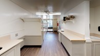 This single-floor laundry facility is designed for practicality and efficiency, offering a clean and well-maintained environment for its users. The space is laid out in a long, open floor plan with light wood-style flooring throughout, contributing to a bright and tidy atmosphere.

Along one side of the facility, a row of white top-load washing machines is neatly aligned, providing ample laundry capacity. Opposite the machines, an extended cream-colored countertop with built-in drop holes runs the length of the room, offering generous surface area for sorting and folding laundry. Louvered cabinet panels beneath the counter provide additional storage and a clean, uniform aesthetic.

The walls are painted in crisp white and feature a decorative shelf adorned with floral arrangements and ornamental figurines, adding a welcoming touch to the functional space. Informational postings, framed notices, and posted guidelines are displayed on the walls for user reference. A wall clock is also visible, keeping the space practical.

Ceiling-mounted fluorescent light fixtures provide consistent, even illumination throughout, while ceiling fans promote comfortable air circulation. Natural light enters through multiple windows, including a larger window at the far end that provides an outdoor view. The overall ambiance is clean, organized, and user-friendly.