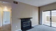 This single-floor residence offers a clean and functional layout designed for comfortable everyday living. The living area serves as the heart of the space, anchored by a standout fireplace clad in dark granite-style tile with a classic wood mantel, adding both warmth and visual interest to the room. The walls are finished in a neutral beige tone, complementing the plush wall-to-wall carpeting that runs throughout the living space and contributes to a cozy, understated atmosphere. Natural light filters into the room through a sliding glass door fitted with vertical blinds, providing a connection to the outdoors while maintaining privacy. Ceiling-mounted HVAC vents are integrated throughout, ensuring year-round climate comfort. The overall aesthetic is simple and neutral, offering a versatile canvas well-suited for a range of interior styles. The single-floor layout promotes ease of movement and accessibility throughout the home. With its functional design, key amenities, and inviting fireplace focal point, this space presents a practical and comfortable living environment.