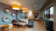 Located in Portland's dynamic Lloyd District, this single-floor amenity lounge at 2881–2889 NE Holladay St offers a thoughtfully designed communal space that balances playful energy with functional versatility. The room is anchored by striking oversized orange drum pendant lights that cast a warm glow across the herringbone-patterned tile flooring, which transitions seamlessly between seating and activity zones. A central low-profile divider separates the lounge seating area from the rest of the room, where a woven bench, upholstered daybed-style seating, and modular ottomans invite relaxed gathering. The accent walls, painted in deep slate blue, are adorned with curated artwork, decorative mirrors, and wire display grids showcasing illustrated prints, giving the space an artsy, eclectic character. A teal bar counter with white stools lines the perimeter near the windows, providing a casual workspace or social bar area with natural light filtering in. Large black-framed glass doors open directly to an outdoor patio, enhancing the connection between interior and exterior. Recessed lighting supplements the pendant fixtures throughout. A designated play area with a small teepee and climbing ladder adds a family-friendly dimension to this energetic, well-appointed community lounge.