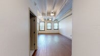 Located at 600 N Stonewall Ave in Oklahoma City, this single-floor unit showcases an industrial-loft aesthetic defined by exposed painted ceiling beams, warm hardwood-style flooring, and crisp white walls throughout. The open-concept kitchen and living area serves as the heart of the unit, where a well-appointed kitchen features dark espresso cabinetry, stainless steel appliances including a refrigerator, dishwasher, range, and microwave, paired with light countertops for a striking contrast. A row of multiple windows along the living area wall floods the space with generous natural light and provides views of the surrounding neighborhood streetscape. The living area offers an open, flexible layout beneath the distinctive exposed beam ceiling, complemented by track lighting that adds an industrial touch. A dedicated entrance leads into the unit, providing a clear transition from the common areas. The walk-in closet offers practical storage with a full-width hanging rod and hardwood-style flooring consistent with the rest of the unit. The bathroom is finished with classic white penny-tile flooring and includes a full tub and shower combination. Overall, the unit delivers a cohesive urban-industrial character with functional, well-considered details throughout.