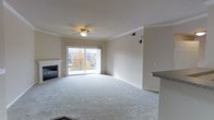 This single-floor unit centers around a spacious, open living area finished with plush wall-to-wall carpeting and neutral-toned walls that create a clean, welcoming atmosphere. Crown molding runs along the ceiling perimeter, adding a subtle architectural detail that elevates the overall aesthetic. A ceiling fan with a light fixture is centrally positioned, providing both air circulation and ambient lighting. The living area includes a fireplace, serving as a functional focal point along one wall. A full-width sliding glass door floods the space with natural light and opens to a private balcony, offering views of the surrounding residential neighborhood, including mature trees displaying autumn foliage. The balcony provides an attractive outdoor extension of the living space. A partial view of an adjacent countertop with granite-style detailing suggests a kitchen area nearby, hinting at a practical open-concept layout. HVAC vents are integrated into the ceiling, and a smoke or carbon monoxide detector is visibly mounted, indicating attentiveness to safety standards. Overall, the space presents a comfortable and functional layout suited for everyday living.