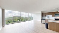 This contemporary 3-bedroom, 2-bathroom residence in Pittsburgh's East End is defined by its striking floor-to-ceiling windows that span the living areas and bedrooms, flooding the space with natural light and framing expansive treetop and skyline views. The open-concept kitchen and living room serve as the heart of the home, where light hardwood flooring flows seamlessly throughout. The well-appointed kitchen features warm wood cabinetry, a granite-topped island with pendant lighting, stainless steel appliances, and a classic subway tile backsplash. The generously sized primary bedroom offers plush carpeting and ample closet space, while the additional bedrooms include a mix of hardwood and carpet flooring, each benefiting from large windows with scenic outdoor views. The two bathrooms are finished with granite countertops, wood-toned vanities, and clean white fixtures. A connecting hallway ties the three bedrooms together, and the unit boasts an impressive total of eight closets, providing exceptional storage throughout. The overall aesthetic is modern and clean, with a neutral palette that enhances the sense of openness and light across every room.