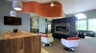 This contemporary single-floor space makes an immediate impression with its bold and thoughtfully designed interior. The most eye-catching architectural feature is the sweeping, wave-shaped ceiling accent finished in a rich terracotta-orange hue, which adds dramatic flair and a sense of movement to the open layout. A ceiling fan is integrated seamlessly into this curved canopy, complementing the modern aesthetic while ensuring comfort.

The space is anchored by a sleek, linear gas fireplace set into a floor-to-ceiling dark cabinetry wall accented with mosaic tile surroundings, creating a sophisticated focal point. Warm wood-toned flooring runs throughout, grounding the vibrant design elements with natural warmth.

Stylish seating — featuring contemporary chairs with bold orange upholstery and polished chrome pedestal bases — adds both function and personality. A substantial wood-topped counter or island adds to the space's versatility, suggesting a lounge, reception, or collaborative area.

A large-scale nature-themed wall mural featuring birch trees brings an organic contrast to the modern design language. Pendant lighting and recessed fixtures contribute to a layered lighting scheme, while glimpses of stainless steel appliances hint at a kitchen or kitchenette tucked nearby. Overall, the space strikes a confident balance between modern sophistication and inviting warmth.