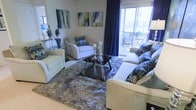 This single-floor space showcases a thoughtfully designed living area that blends comfort with contemporary style. The sitting area serves as the focal point, featuring a neutral-toned sofa and armchairs accented with bold blue and teal patterned throw pillows, creating a cohesive and inviting color palette. A sleek black glass coffee table anchors the seating arrangement, complemented by a plush, textured area rug that adds warmth and depth to the space.

Natural light flows generously into the room through a large sliding glass door dressed with dramatic navy blue floor-to-ceiling curtains, which add a touch of elegance while framing the view to the outdoor balcony. The balcony, visible through the glass, features seating and offers outdoor living potential. Abstract artwork in coordinating blue and green tones adorns the walls, reinforcing the modern aesthetic. Decorative candle holders and sculptural accents add personality to the space. The overall ambiance is polished and welcoming, with a carefully curated design that balances bold decorative choices with a neutral architectural backdrop.