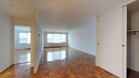 This single-floor space presents a clean, open layout with a notably spacious main living area. The room is defined by its polished parquet hardwood flooring, arranged in a classic block pattern that adds visual texture and warmth to the otherwise neutral interior. The high-gloss finish on the floors reflects light effectively throughout the space.

The walls are finished in crisp white, contributing to a bright and airy atmosphere. A large window positioned along the far wall allows generous natural light to fill the room, with a radiator unit situated beneath it — a functional heating feature typical of well-established residential buildings. The window provides an outward view and serves as the room's primary light source.

The ceiling is smooth and white, maintaining a consistent, unadorned finish that keeps the space feeling open. A closet door is visible along the right wall, offering built-in storage. The layout is flexible and largely open, providing ample room for various furniture arrangements. Overall, the space offers a practical, light-filled environment with classic flooring details and a straightforward, functional design.