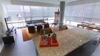 This single-floor space showcases an open-concept layout that seamlessly connects the kitchen and living areas, creating a generous, flowing interior. The kitchen features a substantial granite-topped island with distinctive veining, providing both a functional workspace and a natural focal point. A decorative orange tray with glass bottles and metallic accessories adds a stylish touch to the countertop.

The living area is anchored by a tufted gray sofa accented with patterned throw pillows, complemented by dark cube-shaped coffee tables and an accent chair with a geometric frame. A warm-toned area rug grounds the seating arrangement, while a bold piece of artwork adds a splash of color against the neutral backdrop.

A prominent structural column punctuates the space, adding an architectural element that reinforces the building's character. The most striking feature is the expansive wraparound floor-to-ceiling windows fitted with sheer roller shades, allowing abundant diffused natural light to fill the interior. The overall ambiance is modern and sophisticated, with a carefully curated blend of textures, materials, and warm accent tones throughout.