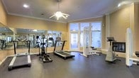This fitness center offers a clean, well-appointed environment designed to support a range of workout routines. The space is outfitted with a selection of cardio machines, including a treadmill and an elliptical trainer, alongside a multi-function cable weight machine, providing users with both aerobic and strength training options. The flooring is finished in a durable dark rubber material, ideal for high-impact exercise and easy maintenance. A full-length mirrored wall runs along one side of the room, effectively doubling the perceived size of the space and allowing users to monitor their form during workouts. Crown molding trims the upper walls, lending a refined, polished character to the interior. A ceiling fan centered overhead provides comfortable air circulation throughout the room. Large frosted-glass French doors and windows along the far wall allow soft, diffused natural light to filter in, creating a bright and inviting atmosphere while maintaining privacy. The warm, neutral wall tones complement the overall aesthetic, striking a balance between functional fitness facility and an elevated, welcoming environment. The layout is thoughtfully arranged to allow for comfortable movement between equipment.