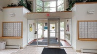 This single-story commercial building makes an immediate impression through its grand entrance lobby, which showcases a double-height ceiling that allows natural light to pour in generously through a large, multi-pane clerestory window. The glazed entryway features floor-to-ceiling glass panels flanking a central glass door, offering unobstructed views of the surrounding outdoor greenery and parking area beyond. Lush potted plants positioned at the upper level of the lobby add a vibrant, natural element to the architectural design, softening the clean, modern lines of the space. The interior finishes are crisp and professional, with light-toned tile flooring accented by a warm terracotta border detail near the entry threshold. Entrance mats are thoughtfully placed at both the exterior and interior sides of the main door, reflecting practical attention to the flow of foot traffic. The walls are finished in a neutral white, complementing the bright, airy atmosphere created by the abundant natural light. Overall, the entrance lobby conveys a welcoming, well-maintained commercial environment designed to make a strong first impression while providing a functional and accessible point of entry.