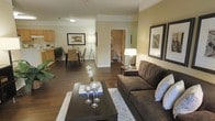 This single-floor residence offers a thoughtfully designed open-concept layout that seamlessly connects the living, dining, and kitchen areas into one cohesive, flowing space. Rich hardwood flooring extends throughout, providing a warm and inviting foundation for the entire floor plan.

The living area is generously sized, accommodating a large sectional sofa, a dark-wood coffee table, and decorative accents including framed wall art and elegant glass centerpieces. Warm ambient lighting from stylish floor lamps enhances the comfortable atmosphere of the space.

The dining area transitions naturally from the living room, featuring a classic dining table with seating for multiple guests, accompanied by tasteful wall art and a potted plant that adds a touch of greenery. Adjacent to the dining space, the kitchen is visible with light wood cabinetry and functional counter space, offering an efficient and accessible cooking environment.

A hallway visible from the main living area leads to additional rooms, including a bathroom, suggesting a practical and well-organized floor plan. The neutral color palette of soft greens and creams on the walls complements the dark furnishings, creating a balanced and harmonious aesthetic throughout the residence.