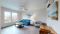 This single-floor unit offers a clean, contemporary design characterized by light-toned wood-style flooring, white walls, and a neutral palette that creates a bright, cohesive atmosphere throughout. The living area serves as the central gathering space, furnished with a blue sofa, a gray accent chair, and a rustic wood coffee table. A ceiling fan with integrated lighting overhead and a large window allow natural light to fill the room, while framed artwork adds a decorative touch. The dining area sits adjacent to the living space, accommodating a wooden table and chairs for casual meals.

The kitchen is efficiently laid out with white cabinetry extending to the ceiling, a full suite of stainless steel appliances including a refrigerator, electric range, and microwave, and a clean white countertop with ample workspace. Recessed lighting illuminates the kitchen for practical use.

The bedroom is a serene retreat featuring large windows that frame views of the surrounding urban and natural landscape, a ceiling fan, and wood-style flooring. Natural light flows freely through the expansive windows, enhancing the sense of openness.

The bathroom includes a shower/tub combination, a single vanity with storage cabinetry, and wood-style flooring. In-unit laundry is provided via a stacked washer and dryer conveniently located nearby.