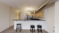 This single-floor space showcases a thoughtfully designed open-concept layout that maximizes its elongated footprint. The kitchen serves as a central highlight, featuring light wood cabinetry extending to the upper walls, dark granite countertops, and a full suite of stainless steel appliances, including a refrigerator, range, and built-in microwave. A wide breakfast bar with seating for three stools bridges the kitchen and the adjoining living area, making it an ideal space for casual dining and entertaining.

A standout architectural detail is the large exposed industrial ductwork running along the ceiling, lending the space a loft-like character. A decorative chandelier pendant light adds a contrasting touch of warmth above the kitchen area. The flooring transitions from carpet in the living areas to hard surfaces near the kitchen, providing both comfort and practicality.

The floor plan, as viewed from above, reveals a narrow yet well-organized layout with clearly defined zones for living, dining, and cooking. Private rooms are positioned toward the rear, separated from the main living areas. The overall aesthetic blends functional design with industrial-inspired elements, creating a balanced and distinctive interior environment.