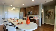 This thoughtfully designed kitchen combines functionality with contemporary style, creating an inviting space for cooking and gathering. Rich, warm-toned cabinetry lines the walls from floor to ceiling, offering generous storage through a combination of upper and lower cabinets. Stainless steel appliances, including a French door refrigerator, a built-in microwave, and a smooth-top range, lend a sleek, modern character to the space.

A standout feature of the kitchen is the expansive white quartz-topped breakfast bar, which extends outward to comfortably seat multiple guests. Its clean, bright surface contrasts beautifully with the dark wood cabinetry and warm hardwood flooring beneath. A stainless steel undermount sink with a modern arc faucet is conveniently integrated into the bar area.

Overhead, recessed lighting is complemented by a chic crystal pendant light fixture that adds an elegant touch above the bar seating. The light sage-green walls contribute a soft, airy ambiance throughout. A glimpse into the adjacent hallway suggests additional living areas beyond. Overall, the kitchen exudes a warm yet polished atmosphere, balancing practicality with refined design details suited for comfortable everyday living.