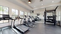 This fitness center is a thoughtfully designed, single-room facility that provides a comprehensive workout environment within a compact yet efficiently utilized footprint. The space is defined by its clean, modern aesthetic, featuring crisp white walls, a tray ceiling with recessed lighting, and a ceiling fan that contributes to air circulation during workouts. Dark, durable rubberized flooring runs throughout, providing both safety and practicality underfoot.

Natural light floods the room through a series of large, black-framed windows along one wall, offering views of the surrounding exterior and creating a bright, energizing atmosphere. A full-length mirror along one wall visually expands the space while allowing users to monitor their form during exercise.

The cardio section is well-stocked, featuring multiple treadmills, elliptical trainers, and a stationary spin bike. A rowing machine and a multi-function cable strength machine provide additional training variety. Free weights on a dedicated rack, a heavy punching bag, and a wall-mounted television round out the amenities. Entry is provided through glass-paneled double doors, and a keypad access system ensures secure entry. Fitness center rules are posted on the wall, reinforcing a structured, community-oriented environment.