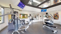 Located at 8115 Worthington Galena Rd in Westerville, Ohio, this fitness center at Verve is a thoughtfully designed exercise facility offering a comprehensive workout environment. The space is defined by its striking architectural ceiling feature — a raised, dark-framed skylight structure fitted with mirrored panels that amplify the sense of height and openness while flooding the room with natural light. Multiple windows along the walls further enhance brightness and provide views of the surrounding greenery.

The exercise room is outfitted with a wide array of equipment to support both cardio and strength training. Cardio offerings include treadmills, elliptical machines, a stationary bike, and a rowing machine. For strength training, the space features a multi-station cable machine, adjustable benches, and a full dumbbell rack. A dedicated functional training area along one wall includes medicine balls, stability balls, and a suspension training system, complemented by motivational fitness-themed wall art.

Multiple flat-screen TVs are wall- and ceiling-mounted throughout the space, ensuring entertainment is visible from virtually any workout station. The flooring consists of durable, dark rubber gym matting, and the clean white walls contribute to a bright, modern aesthetic. Full-length mirrors along one wall enhance spatial depth and allow users to monitor their form during exercise.
