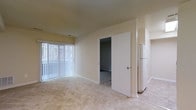 Unit 119 is a single-story space finished in a clean, neutral palette that creates a bright and welcoming atmosphere throughout. The main living area is an open, carpeted room with soft beige walls and a large window dressed with vertical blinds, allowing natural light to filter in while maintaining privacy. The light-toned carpet continues through the space, adding warmth underfoot.

A wide doorway leads to an adjacent room, offering flexible use as a bedroom or additional living area, also finished with matching carpet and neutral walls. To the right, a partial view reveals a white refrigerator, indicating the presence of a kitchen or kitchenette nearby. Clean white trim and door frames provide a crisp contrast against the warm wall tones throughout.

The overall layout is straightforward and practical, with a simple, uncluttered design that allows residents to personalize the space with ease. The consistent neutral finishes across all visible areas give the unit a cohesive, move-in-ready feel, while the natural light from the main window enhances the sense of openness in the living space.