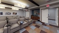 This single-floor media and screening room is thoughtfully designed to deliver a cinematic experience in a comfortable, lounge-style setting. The focal point of the space is a large wall-mounted flat-screen television positioned against a textured, dark accent wall, flanked by a dark wood media console with cabinet storage below. Rows of plush, light gray upholstered armchairs are arranged in a tiered, theater-inspired layout, each equipped with a personal fold-out tablet tray for added convenience. The flooring features a bold, patchwork-style carpet in earthy tones of brown, tan, gray, and rust, adding visual interest and warmth throughout. Decorative dark wood ceiling beams run overhead, complemented by recessed lighting and large pendant fixtures that provide ample, adjustable illumination. One side of the room is lined with floor-to-ceiling drapes in a rich navy and gold pattern, offering light control and an elegant finish. Along the back wall, a built-in wood cabinetry unit with a dark countertop serves as a refreshment or service station. Contemporary framed artwork adorns the walls, and decorative vases with floral arrangements add tasteful accents. The overall ambiance is modern, polished, and well-suited for private screenings or group entertainment gatherings.