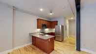 This single-floor space centers around a thoughtfully designed kitchen that combines functionality with a warm aesthetic. The kitchen features rich cherry wood cabinetry that extends to the upper walls, offering generous storage throughout. A peninsula island with a granite countertop serves as the focal point, complete with an integrated sink and additional seating potential, making it ideal for both meal preparation and casual dining.

Stainless steel appliances, including a full-size refrigerator, built-in microwave, and range, lend a modern, cohesive look to the space. A classic subway tile backsplash adds texture and contrast against the warm wood tones. Light hardwood flooring runs throughout, brightening the interior and providing a seamless flow between areas. Recessed lighting and track lighting ensure the kitchen and adjacent spaces are well-illuminated at all times.

The layout opens naturally into a hallway leading to additional areas of the unit, suggesting a practical and well-organized floor plan. Neutral gray walls complement the warm wood and light flooring, creating a balanced and inviting overall ambiance.