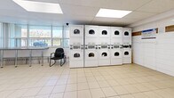 Located at 20 Esterbrooke Ave in Toronto's M2J neighbourhood, this shared laundry facility is a well-organized and functional communal space designed to serve building residents efficiently. The room features light-toned ceramic tile flooring throughout and a suspended drop ceiling fitted with recessed panel lighting, creating a bright and clean environment.

The space is generously equipped with multiple top-loading washing machines along one wall, complemented by a bank of stacked front-loading dryers that maximize vertical space and accommodate a high volume of laundry. Additional front-loading washers are also present, offering residents a variety of machine options. A utility sink provides added convenience for hand-washing or pre-treating garments.

A long stainless steel folding counter runs along the windowed wall, offering ample surface area for sorting and folding laundry. Large windows along this wall flood the space with natural light and provide views of the surrounding greenery and outdoor area, lending an open and airy feel. Seating is available near the windows, allowing residents to wait comfortably.

The walls are finished with white tile accented by a decorative horizontal band of tan tile, contributing to the clean, utilitarian aesthetic. Informational signage is posted throughout, and a waste bin is conveniently placed. The space is accessed through a dark-painted door and is monitored by a security camera.