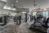 This fitness center is a comprehensively outfitted facility designed to support a full range of workout routines. The space is organized into distinct functional zones, allowing users to move seamlessly between cardio training, strength work, and functional fitness exercises. A row of True-brand treadmills and ellipticals lines one side of the room, each equipped with individual screens for entertainment during workouts. Large floor-to-ceiling mirrors span an entire wall, visually expanding the space and allowing users to monitor their form. The free weight section features a full dumbbell rack alongside adjustable benches, cable machines, and various strength training apparatus. A dedicated functional training area includes medicine balls, resistance equipment, and a cable crossover station. Multiple wall-mounted flat-screen televisions are positioned throughout the room for convenient viewing. Auburn University branding is prominently displayed, adding a collegiate character to the environment. The floor is covered in a patterned, multi-toned modular carpet that defines different workout zones with subtle color accents. Elevated ceilings with recessed lighting and pendant fixtures, combined with large windows offering outdoor views, create a bright and energizing atmosphere throughout the facility.