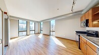 This one-bedroom, one-bathroom urban residence offers a distinctive industrial-loft aesthetic across a single thoughtfully laid-out floor. The open-concept living and kitchen area is defined by striking floor-to-ceiling windows on multiple walls, flooding the space with abundant natural light and framing views of the surrounding cityscape and neighboring architecture. Exposed metallic ductwork runs along the ceiling, adding an industrial character that complements the light hardwood flooring found throughout the main living areas. The kitchen is equipped with warm wood cabinetry, dark countertops, and a full suite of appliances including a stainless range, built-in microwave, dishwasher, and refrigerator — all anchored by track lighting overhead. The bedroom is a comfortable, carpeted retreat featuring a double sliding-door closet, a large window with exterior views, and the same exposed duct detailing as the rest of the unit. Hallways connect the main living areas efficiently, with a utility panel and thermostat control accessible along the way. The bathroom features a full soaking tub with shower, white surround, tile flooring, and a wood-accented vanity with a clean white countertop. The overall ambiance is bright, contemporary, and well-suited for urban living.