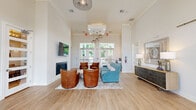 This single-floor entrance and lounge space offers an open, airy atmosphere defined by soaring ceilings and crisp white walls that create a bright, welcoming ambiance. Light-toned wood flooring runs throughout, complementing the clean, contemporary design aesthetic. The main lounge area is centered around a sleek linear fireplace topped with a wall-mounted television, flanked by rich cognac leather swivel chairs and a teal upholstered sofa arranged atop a soft area rug. Stylish pendant lighting and a sculptural wire chandelier add visual interest overhead. Large glass-paneled double entry doors flood the space with natural light and provide views of the surrounding greenery outside. A built-in wet bar or refreshment station features dark wood cabinetry, a granite countertop, and a beverage refrigerator beneath an arched decorative mirror—ideal for welcoming guests. A console table with table lamps and a framed mirror enhances the reception area's refined feel. Glass-paneled interior French doors lead to what appears to be a private meeting or conference room, maintaining visual openness while offering separation. The overall design blends modern comfort with sophisticated detailing, making this entrance a polished and functional gathering space.