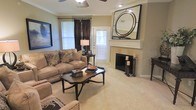 This single-floor residence offers a thoughtfully designed living space that balances comfort and elegance. The living room serves as the heart of the home, centered around a classic fireplace with a clean white mantel, above which a striking large circular mirror adds a bold decorative focal point. Plush wall-to-wall carpeting in a soft neutral tone runs throughout, contributing to the warm and welcoming atmosphere. The space is furnished with a generously sized beige sofa and coordinating seating, complemented by a dark ornate coffee table that adds contrast and character. Crown molding along the ceiling elevates the architectural detail, while a ceiling fan with integrated lighting provides both practical airflow and ambient illumination. Natural light filters into the room through multiple windows dressed with rich, dark curtain panels, offering a refined visual balance between light and warmth. A door with shuttered sidelights provides additional brightness and hints at direct access to the exterior. The neutral wall tones and carefully chosen furnishings create a cohesive, inviting environment throughout. Overall, this residence presents a polished and comfortable living experience with classic design elements that lend it a timeless appeal.