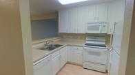 This single-floor space centers around a well-organized kitchen that showcases a cohesive, clean aesthetic. The kitchen is outfitted with white cabinetry that extends to the upper walls, providing generous storage capacity through a combination of upper and lower cabinets. The countertops feature a neutral, light-toned laminate surface that runs along an L-shaped layout, maximizing workspace efficiency. A stainless steel double-basin sink is integrated into the countertop and paired with a chrome faucet, positioned beneath a pass-through opening that connects to an adjacent area, adding a sense of openness to the layout. White appliances, including an electric range with overhead microwave and a dishwasher, are seamlessly incorporated into the cabinetry design, maintaining a uniform and tidy appearance. Light hardwood-style flooring runs throughout, complementing the neutral wall tones and bright cabinetry. Overhead, a flush skylight-style ceiling fixture provides broad, even illumination across the workspace. The overall ambiance is practical and clean, with a straightforward design suited for everyday use.