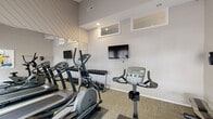 This fitness center is housed within a single-story space distinguished by vaulted ceilings that lend a sense of openness and airiness to the room. The elevated ceiling height is accentuated by angled shiplap detailing, contributing a clean, modern aesthetic to the overall design. Neutral gray walls and patterned carpet flooring provide a polished yet functional backdrop throughout.

The facility is well-stocked with cardio equipment, including multiple treadmills and elliptical trainers positioned along one side of the room, complemented by stationary and upright exercise bikes. A dedicated free weight area features a full rack of dumbbells organized in ascending order. Wall-mounted flat-screen televisions are positioned for easy viewing during workouts.

Floor-to-ceiling mirrors line one wall, visually expanding the space while allowing users to monitor their form. Natural light enters through multiple windows, including elevated clerestory-style windows and standard windows near the entry, keeping the environment bright throughout the day. A glass-paned door provides direct access to an outdoor area, further enhancing the connection to natural light.

A fitness center rules-of-use board and hand sanitizing station are positioned near the entrance, reinforcing a clean and organized environment. The space connects to an adjacent common area, visible through an interior pass-through window.