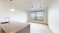 Located at 2528 Texas Ave in St. Louis' vibrant Fox Park neighborhood, this single-floor loft-style unit masterfully blends industrial heritage with contemporary design. Throughout the space, light-toned hardwood flooring provides a warm, cohesive foundation that complements the crisp white walls and soaring ceilings with exposed ductwork — hallmarks of the building's adaptive reuse character.

The open-concept kitchen is a standout feature, anchored by a substantial island with a polished stone countertop in a rich dark hue and a sleek blue base. Stainless steel appliances, including a refrigerator, microwave, and range, are paired with a mix of dark and white cabinetry accented by brushed gold hardware. A gold-toned faucet adds a refined touch, while a decorative tile backsplash and pendant globe lighting complete the sophisticated aesthetic. An ornamental column in the kitchen area adds an architectural nod to the building's history.

The living and sleeping areas are generously proportioned, with oversized, multi-pane black-framed windows that flood the rooms with natural light and frame panoramic views of the surrounding St. Louis cityscape. Exposed brick detailing around the window surrounds further reinforces the loft's industrial character. Dark interior doors with gold hardware provide elegant contrast throughout. The unit offers a clean, versatile canvas well-suited to a variety of uses.