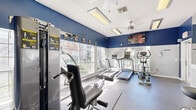 Located at 5500 Sampson St in Houston, Texas, this fitness center is a well-appointed, single-room exercise facility designed to maximize functionality within a compact footprint. The space is defined by its bold two-tone color scheme, featuring rich navy blue upper walls contrasted against crisp white lower walls, creating an energetic and motivating atmosphere.

Large, multi-pane windows line the walls, flooding the room with abundant natural light while offering views of the surrounding outdoor greenery. Full-length mirrors are mounted along one wall, visually expanding the space and allowing users to monitor their form during workouts. The durable rubber flooring provides a safe, slip-resistant surface throughout.

The fitness center is equipped with a comprehensive selection of cardio machines, including treadmills, an elliptical trainer, and an upright stationary bike. A multi-function cable weight machine and an adjustable workout bench are also present, supporting a variety of strength training exercises. Motivational fitness posters are displayed along the upper walls, adding character to the space.

Overhead, flush-mounted fluorescent light panels and a ceiling fan ensure consistent illumination and comfortable air circulation. Wall-mounted flat-screen televisions provide entertainment during workouts. A French door entry and a secondary exit door offer convenient access. The space is also equipped with a fire extinguisher for safety.