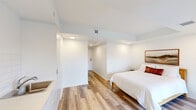 Located at 2376 Dundas St W in Toronto's Junction neighbourhood, this self-contained guest suite offers a thoughtfully designed layout across a single floor. The interior features a clean, contemporary aesthetic defined by bright white walls, recessed pot lighting, and light-toned wood laminate flooring that runs throughout the main living area, contributing to a cohesive and airy feel.

The spacious primary bedroom serves as the suite's central living space, accommodating a bed, seating area with an armchair, and a dedicated workspace with a desk and chair. A framed landscape artwork above the bed adds a warm decorative touch. Large floor-to-ceiling windows and a glass sliding door flood the room with natural light and provide direct access to an outdoor terrace, offering views of the surrounding neighbourhood.

A compact kitchenette with white cabinetry and a sink is integrated into the space, providing basic amenities. The private bathroom is finished in light neutral tones, featuring tile flooring, a vanity with storage, a toilet, and a separate shower stall. A dedicated entrance corridor leads from the suite door into the main living area, providing a clear sense of separation upon entry. Overall, the suite presents a functional and modern environment suited for short-term or guest accommodations.
