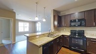 This single-floor space showcases an open-concept design that seamlessly connects the kitchen and living area, creating a cohesive and functional environment. The kitchen is a standout feature, equipped with rich dark espresso cabinetry that provides ample storage, complemented by light-toned granite countertops with subtle gold and beige veining. A peninsula extends the counter space, doubling as a breakfast bar and enhancing the kitchen's practicality. The stainless steel double-basin sink is positioned on the peninsula, paired with a modern arc faucet. Black appliances, including a range and built-in microwave, lend a sleek, consistent aesthetic throughout the kitchen. Three pendant lights with white shades hang above the peninsula, providing focused task lighting while adding visual interest. The open layout flows naturally into the living area, where large windows allow generous natural light to fill the space, contributing to an airy and bright atmosphere. Neutral gray walls and light hardwood-style flooring tie the spaces together, creating a clean and modern ambiance throughout this well-appointed single-floor home.