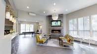 This single-floor space presents a thoughtfully designed communal lounge area that balances comfort and contemporary style. The open-plan layout creates a welcoming atmosphere, anchored by a striking floor-to-ceiling accent wall clad in rustic reclaimed wood, which frames a sleek linear gas fireplace and a wall-mounted flat-screen television. The combination of natural and modern materials gives this focal point a warm yet refined character.

Seating is arranged to encourage conversation, featuring bold yellow upholstered chairs and striped sofas with coordinating accent pillows, all positioned atop a patterned area rug that defines the lounge zone. Slim, minimalist side tables complement the contemporary aesthetic. Recessed ceiling lighting, combined with a prominent pendant light fixture, ensures the space is well-lit throughout.

The tile flooring extends seamlessly across the room, lending a cohesive, polished look. Natural light enters through multiple windows fitted with blinds, adding brightness to the neutral grey and white color palette. In the background, a dedicated game area — featuring what appears to be an air hockey table — provides an additional layer of recreational functionality. A wall-mounted mini-split HVAC unit ensures year-round climate comfort.