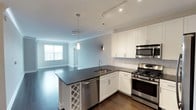 This well-appointed single-floor residence showcases a thoughtfully designed open-concept layout that seamlessly connects the kitchen and living areas. The kitchen is a standout feature, outfitted with crisp white shaker-style cabinetry, dark quartz countertops, and a full suite of stainless steel appliances including a gas range, built-in microwave, dishwasher, and refrigerator. A marble-style tile backsplash adds a refined touch, while pendant lighting and a track lighting system illuminate the space effectively. The kitchen island offers additional prep space and includes a built-in wine rack, making it ideal for entertaining. Rich, dark hardwood flooring flows throughout the main living areas, complementing the neutral wall tones and crown molding details. The bedroom is generously proportioned, featuring the same hardwood floors, track lighting, and large windows that invite natural light. The bathroom is finished with a dark-framed vanity mirror, dual-sink white countertop, dark cabinetry with brushed nickel hardware, and light tile flooring. Together, the finishes and layout create a clean, contemporary ambiance with a strong attention to detail throughout.